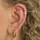 Close-up of a white woman with curly blonde hair wearing a 14K solid gold four-point cluster CZ threadless flat back earring on the helix, paired with a diamond stud on the lobe, showcasing natural skin texture and a curated ear stack in soft lighting