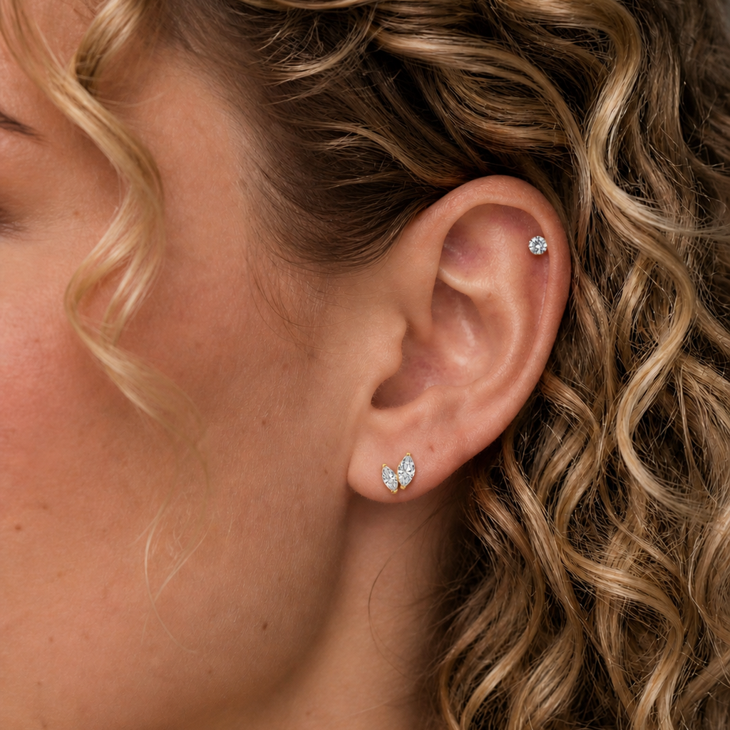 White woman with curly blonde hair wearing DOVIANA graduated double marquise CZ threadless labret titanium PVD gold flat back earring on lobe with diamond stud on helix, natural skin texture close-up