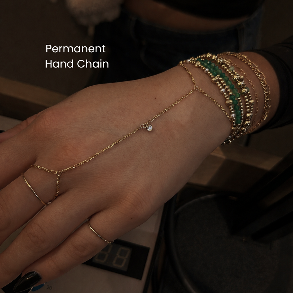 Close-up of a gold permanent hand chain bracelet connected from finger to wrist, layered with multiple gold and green beaded bracelets, showcasing dainty luxury jewelry and permanent hand chain styling in NYC.