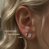 Close-up of a blonde woman’s ear showing DOVIANA stacked lobe and second lobe piercings with dainty diamond stud earrings, illustrating curated ear styling and professional ear piercing placement.