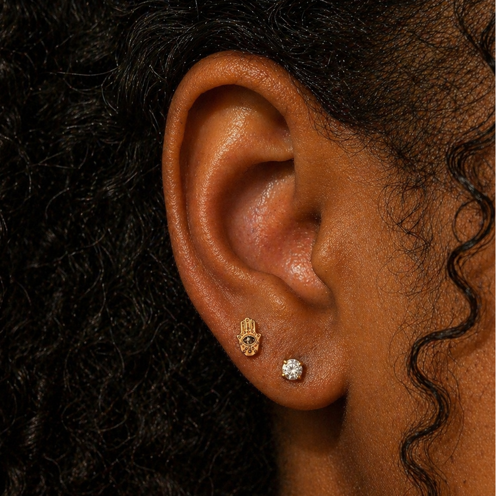 Close-up of ear wearing an evil eye Hamsa hand threadless flat back earring in gold titanium, styled with a diamond stud on the lobe, shown on a Black woman with natural curly hair at DOVIANA NYC