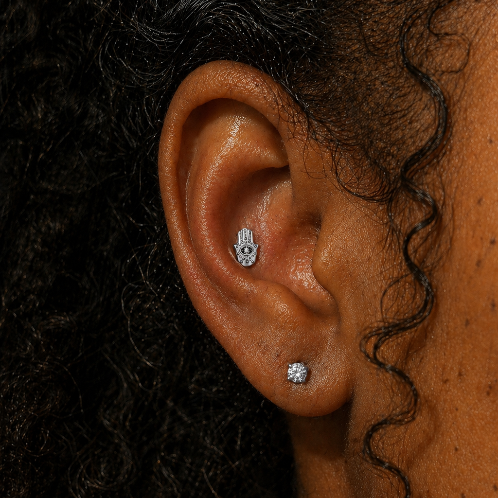 Close-up of ear wearing an evil eye Hamsa hand threadless flat back earring in silver titanium, styled with a diamond stud on the lobe, shown on a Black woman with natural curly hair at DOVIANA NYC