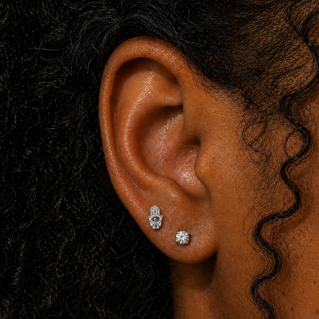 Close-up of ear wearing an evil eye Hamsa hand threadless flat back earring in silver titanium, styled with a diamond stud on the lobe, shown on a Black woman with natural curly hair at DOVIANA NYC
